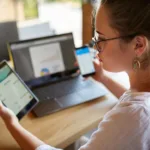 Mixed race woman in glasses working with multiple electronic internet devices. Freelancer businesswoman has tablet and cellphone in hands and laptop on table with charts on screen. Multitasking theme