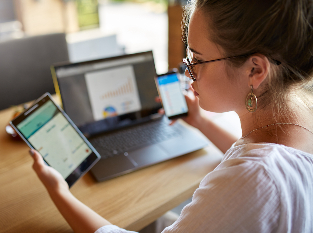 Mixed race woman in glasses working with multiple electronic internet devices. Freelancer businesswoman has tablet and cellphone in hands and laptop on table with charts on screen. Multitasking theme