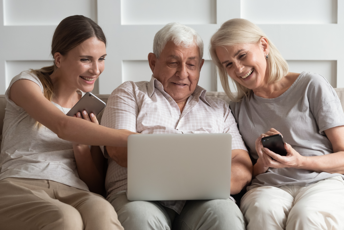 a senior husband and wife sitting on a couch with their adult daughter who is holding a phone all looking at senior living communities on a laptop