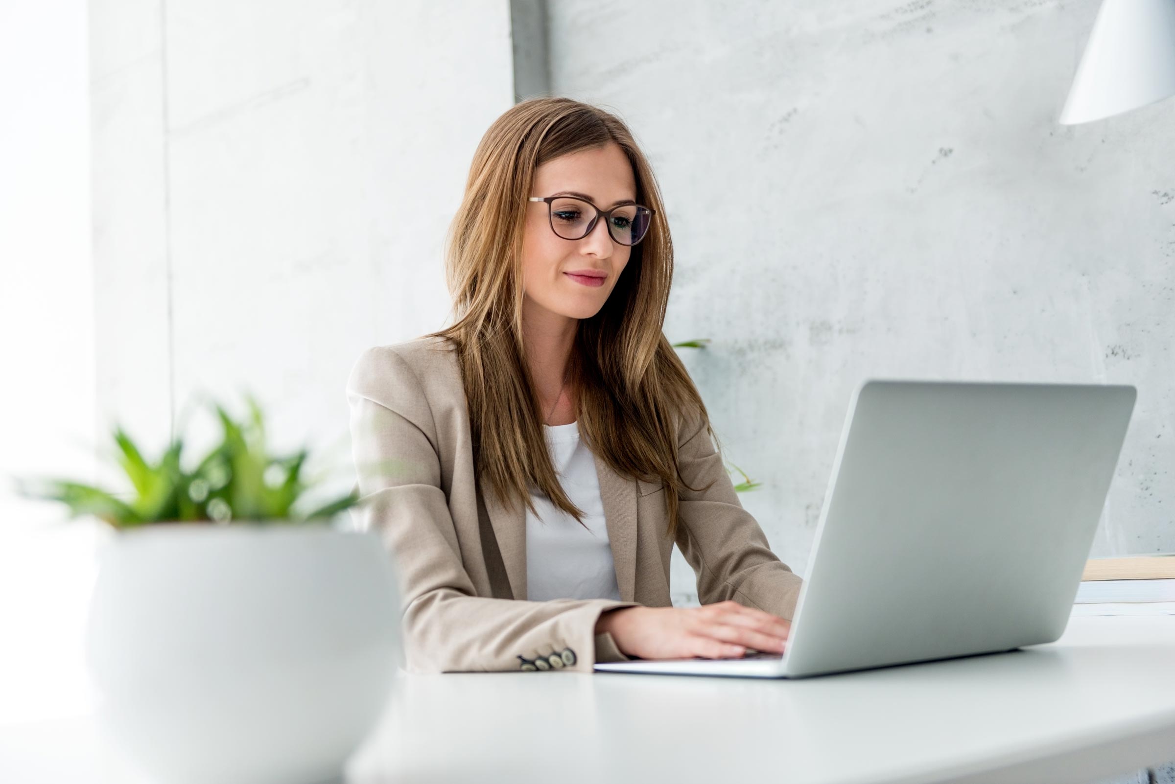 a woman Marketing manager wearing glasses working on her laptop at her desk with a plant on the desk